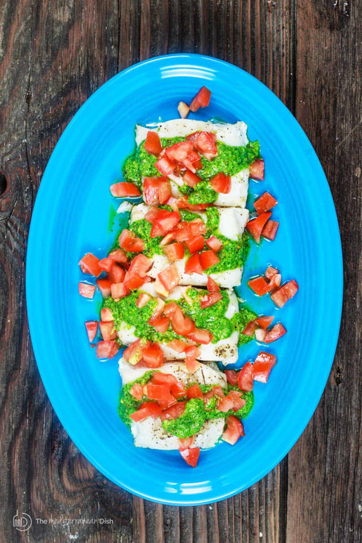 baked cod with pistachio cilantro pesto on a blue serving platter, topped with diced Roma tomatoes