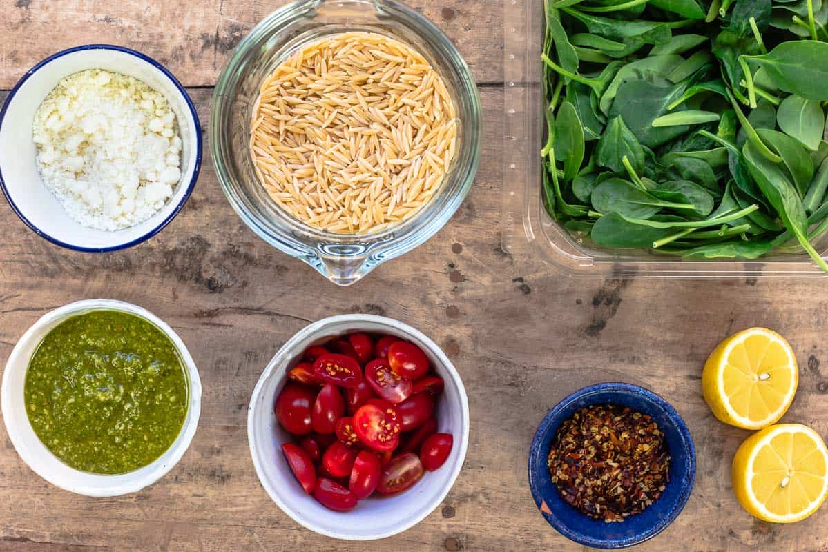 ingredients for orzo pasta soup: fresh lemon, baby spinach, uncooked orzo, grated parmesan, basil pesto, halved cherry tomatoes, red pepper flakes