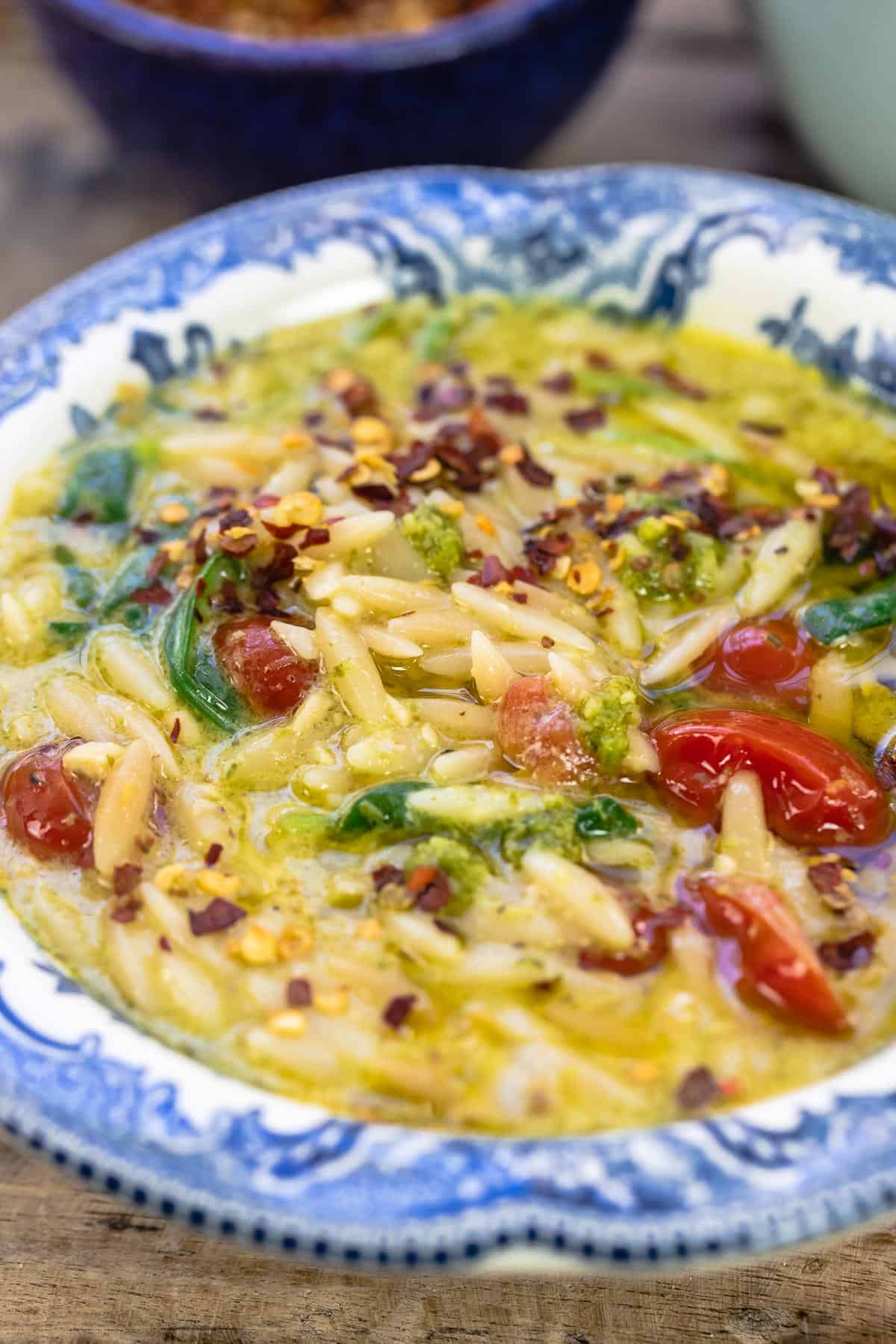 a close up side angle photo of pesto lemon orzo soup in a blue bowl with a side of crushed pepper flakes. 