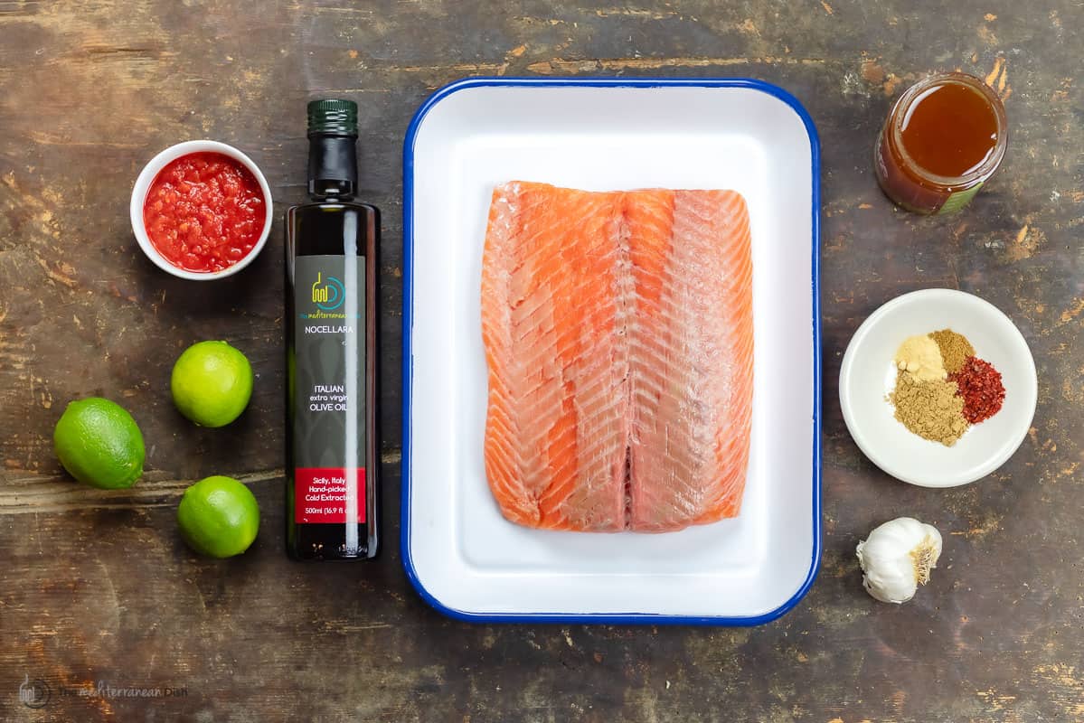 ingredients for spicy salmon including salmon fillet, spices, honey, garlic, limes, extra virgin olive oil, and harissa paste