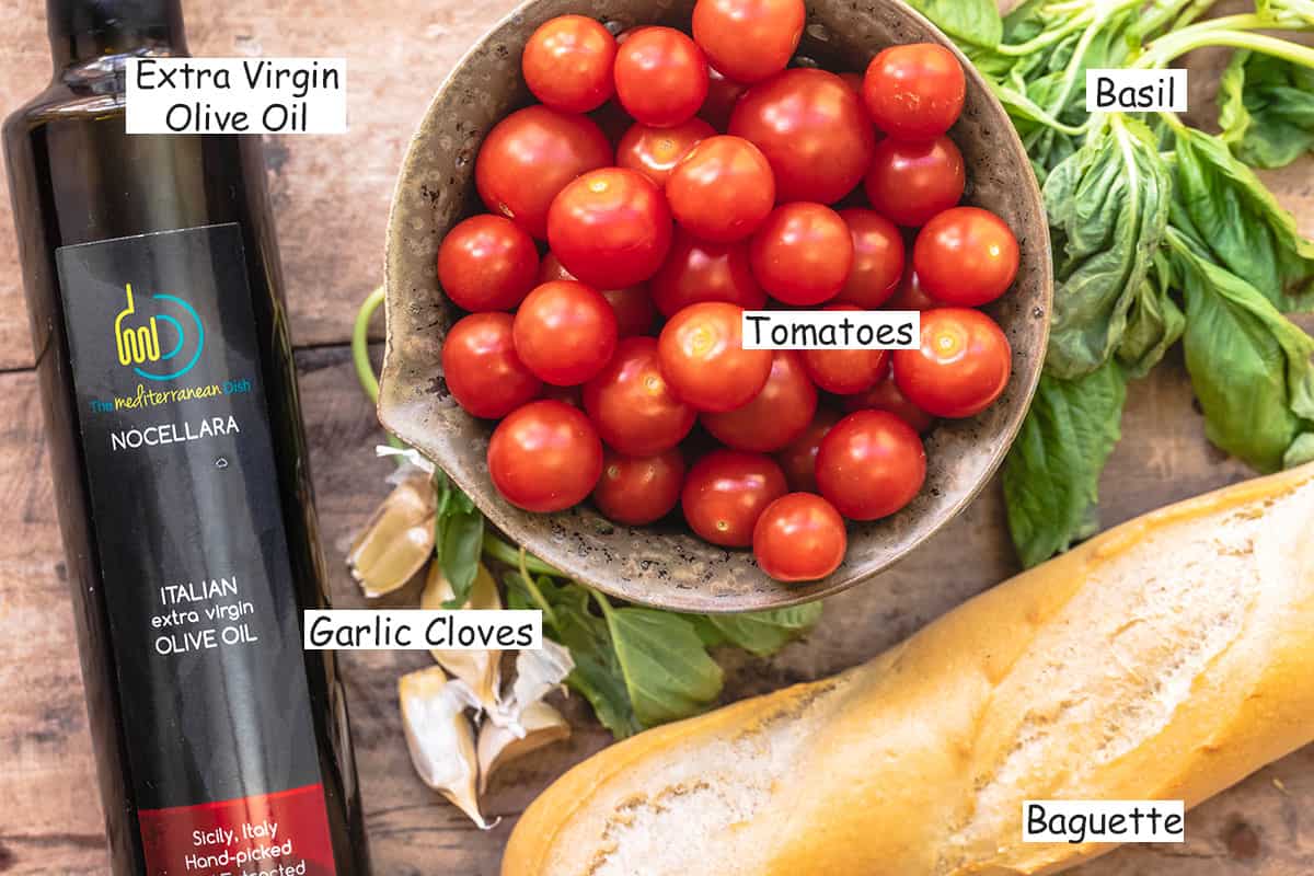 labeled ingredients for tomato bruschetta including extra virgin olive oil, garlic, tomatoes, baguette, and fresh basil