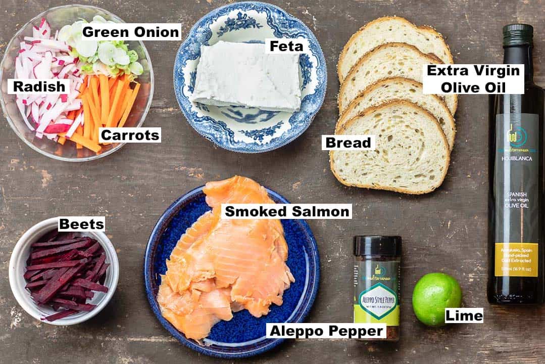labeled ingredients for smoked salmon toast including extra virgin olive oil, bread, feta cheese, vegetables, smoked salmon, Aleppo pepper, and lime