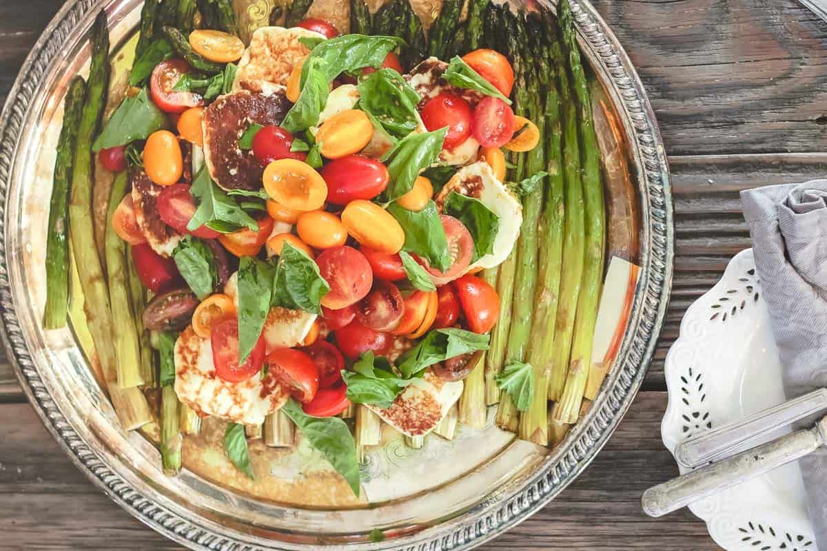 roasted asparagus salad with cherry tomatoes, halloumi cheese, and basil leaves on a platter next to a plate and flatware