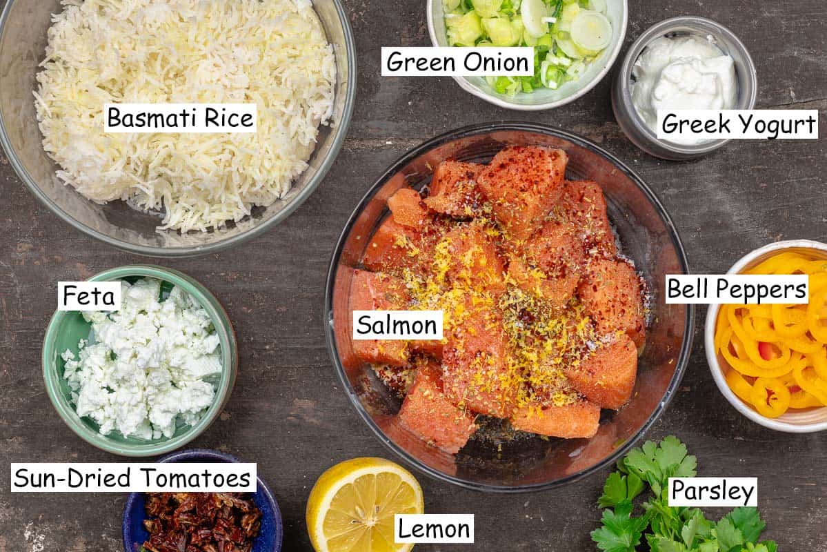 labeled ingredients for salmon rice bowl including cooked basmati rice, green onion, greek yogurt, bell peppers, parsley, seasoned salmon, lemon, sun-dried tomatoes, and feta cheese