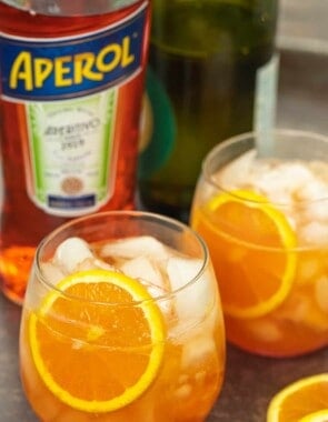 two aperol spritz cocktails with fresh orange slices and a bottle of aperol in the background.