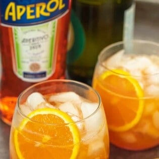 two aperol spritz cocktails with fresh orange slices and a bottle of aperol in the background.