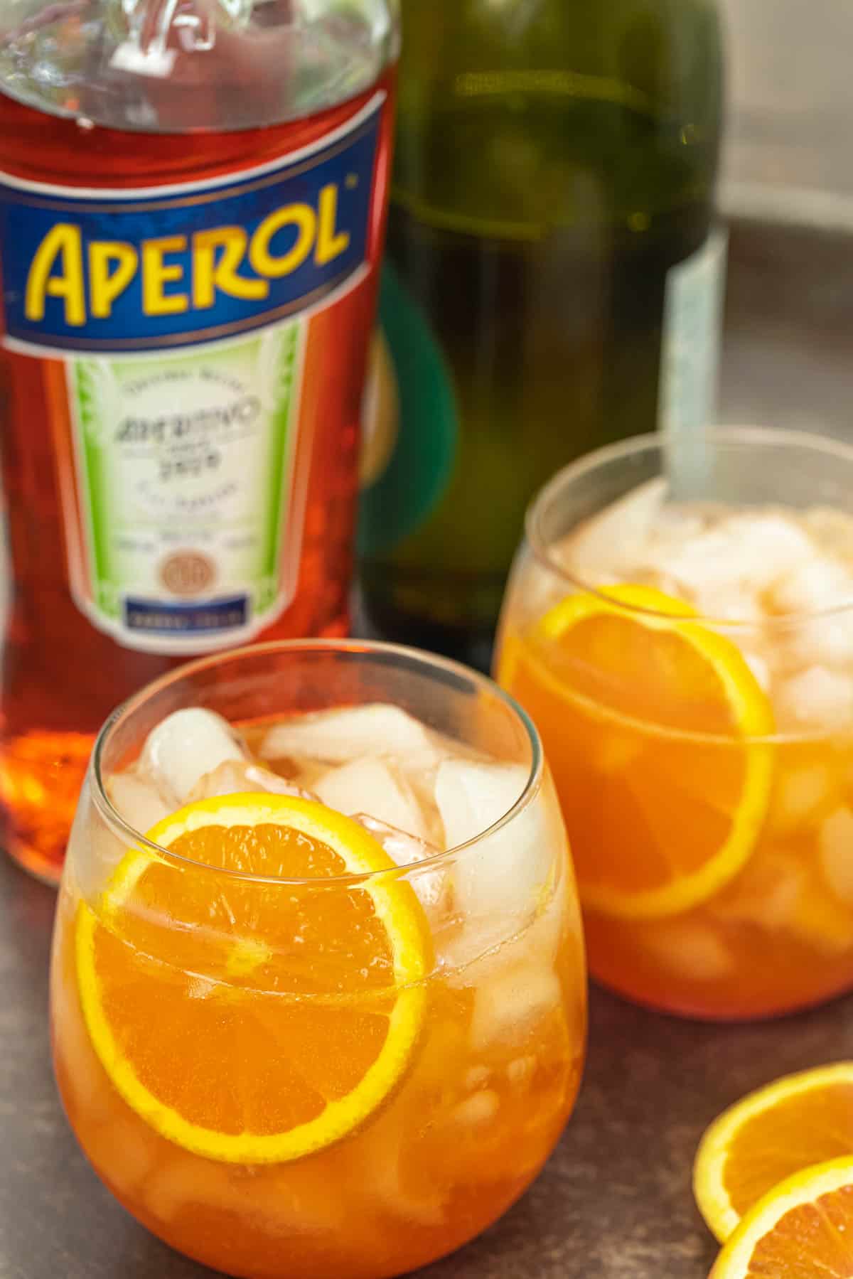 two aperol spritz cocktails with fresh orange slices and a bottle of aperol in the background.
