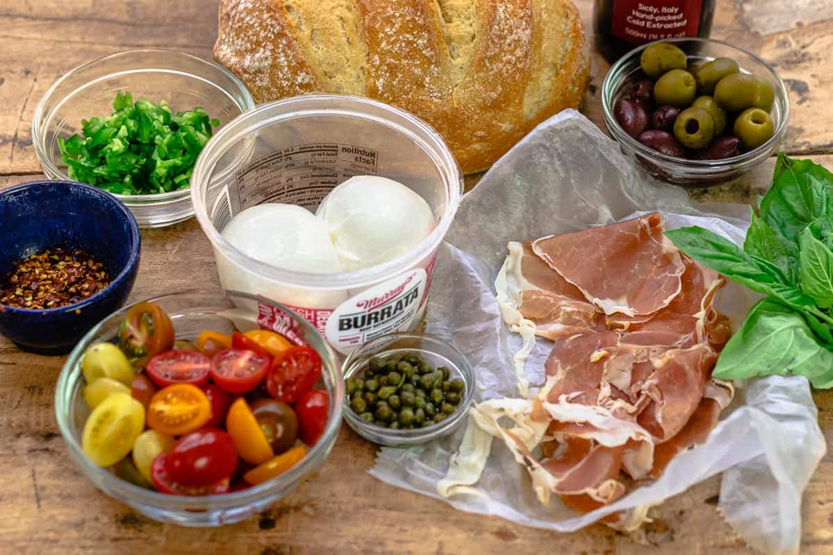 ingredients for burrata recipe including burrata cheese, cherry tomatoes, red pepper flakes, capers, prosciutto, seeded minced jalapeno, ciabatta, olives, basil, and olive oil.