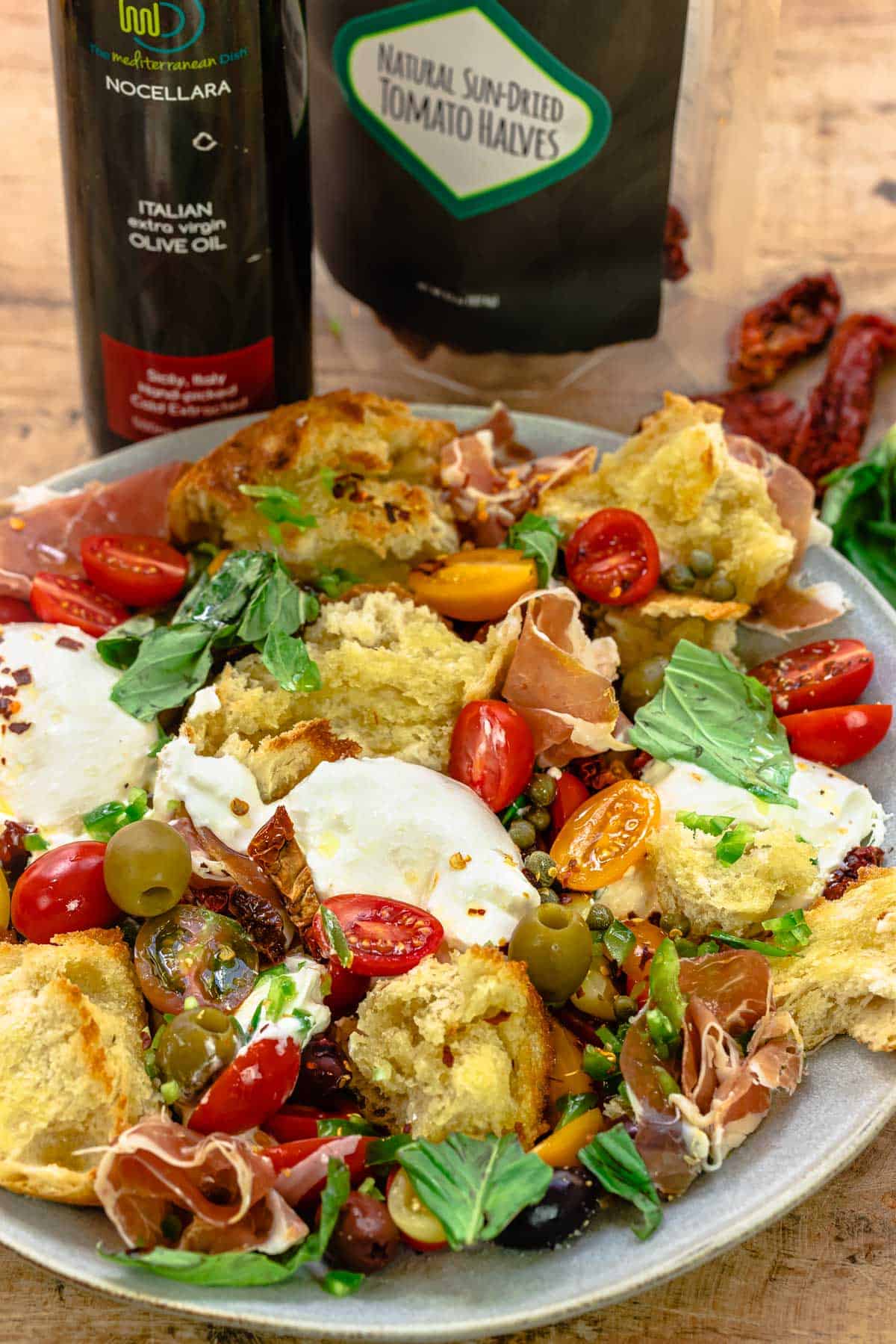 burrata with tomato, basil, and prosciutto and other ingredients on a plate with a bottle olive oil and a bag of sun-dried tomatoes in the background.