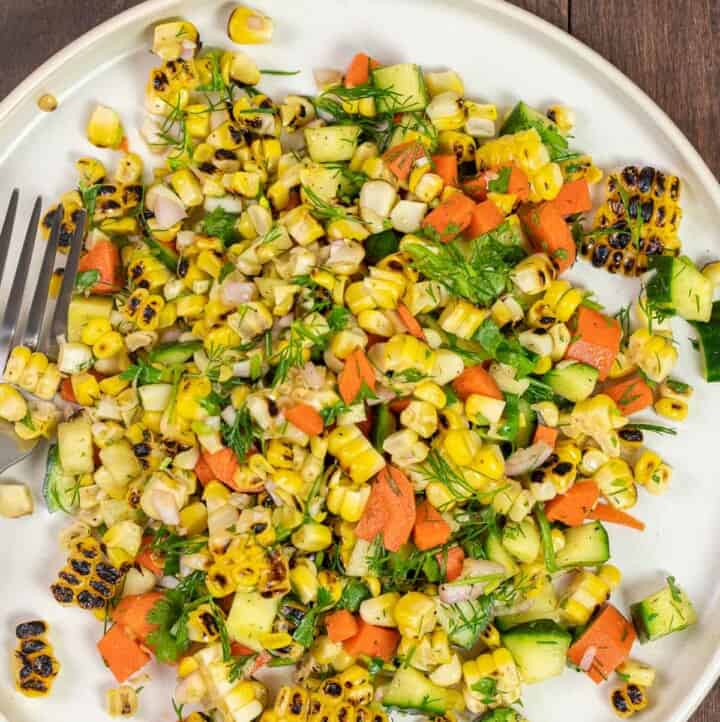 corn salad with cucumber, carrots, and fresh herbs on a white plate with a fork.