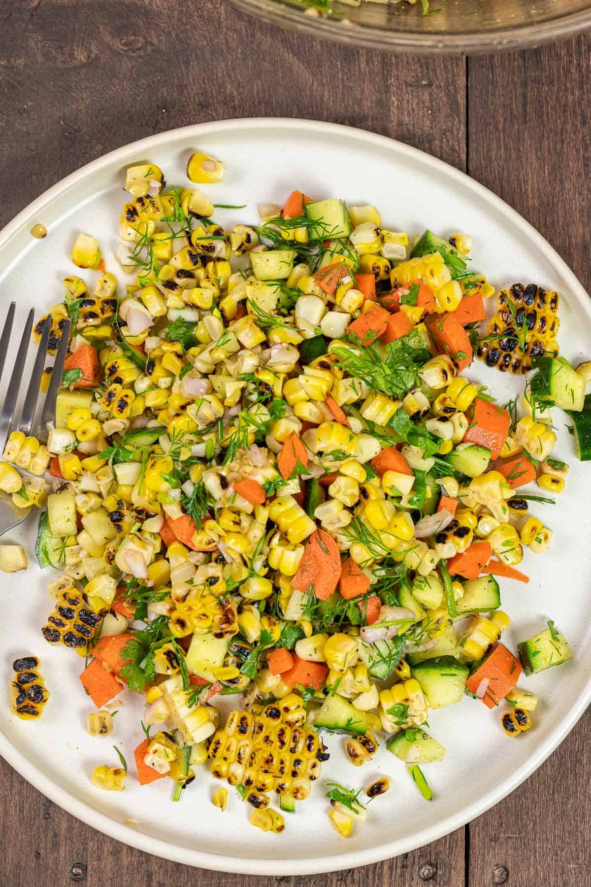 corn salad with cucumber, carrots, and fresh herbs on a white plate with a fork.