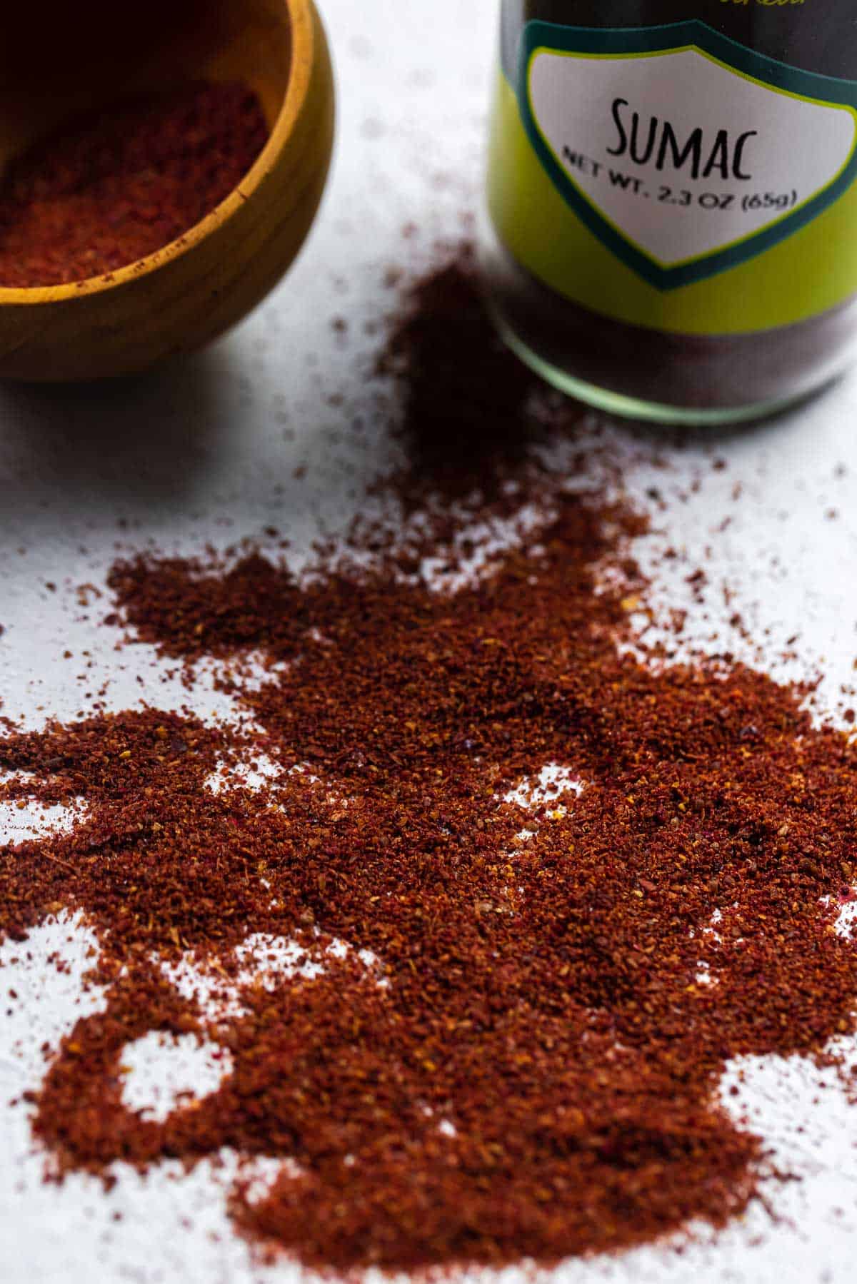 deep red sumac spice sprinkled onto a flat surface with a spoon full of the spice and a bottle of The Mediterranean Dish sumac in the background. 