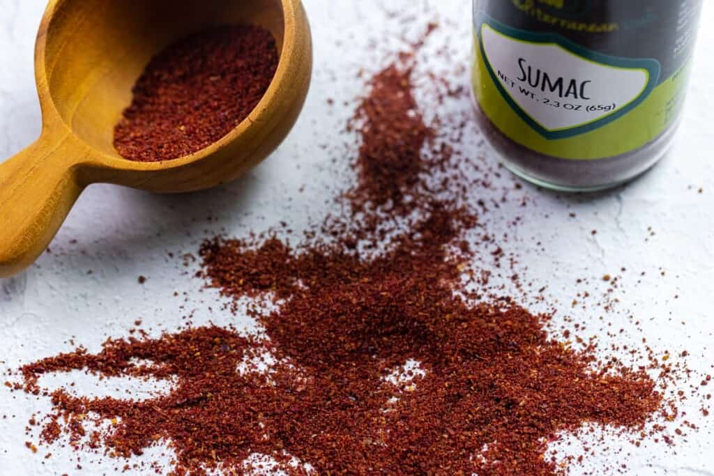 red sumac spice on a white surface with a spoon half filled with the spice and part of The Mediterranean Dish sumac bottle in the back