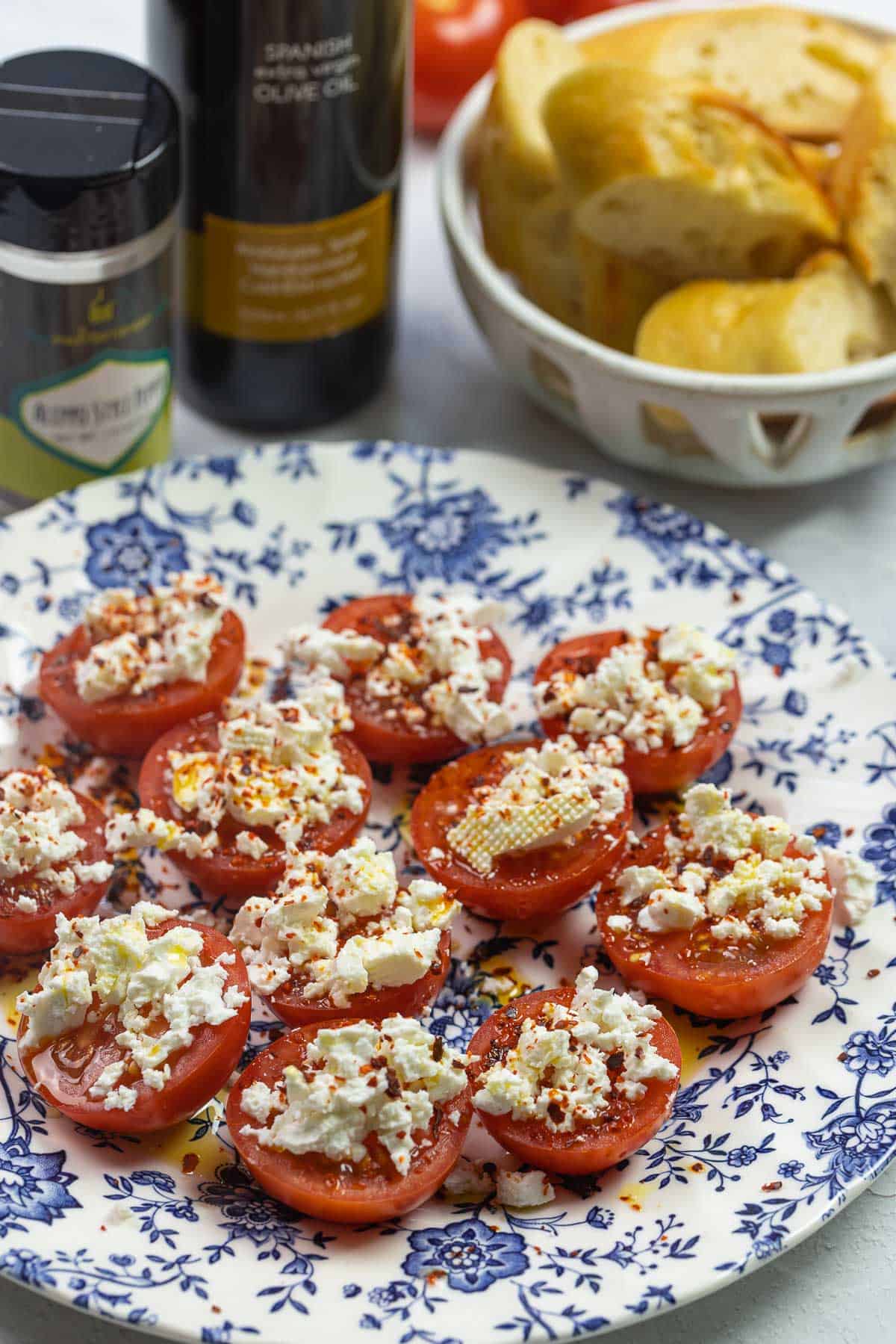 Campari tomato slices with crumbled feta cheese and red pepper flakes. Seasoning bottles to the side. A bread basket to serve along.