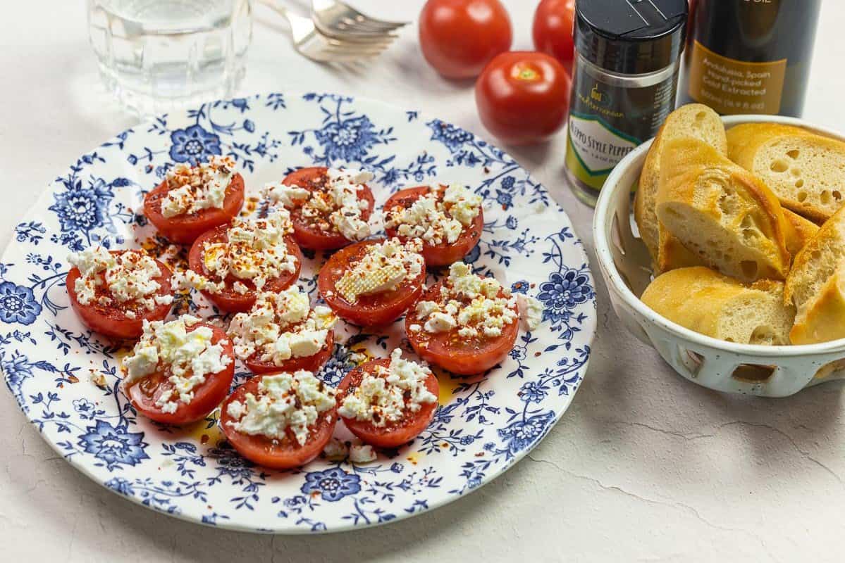 tomato appetizer with sliced tomatoes and crumbled feta and bread basket. A glass of water and bottler of seasoning and an olive oil bottle to the side.