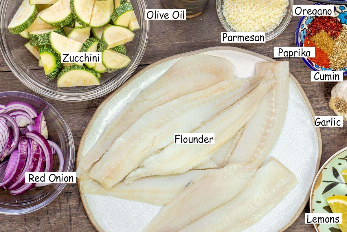 labeled ingredients for baked flounder recipe including flounder fillets, zucchini, red onion, olive oil, parmesan cheese, oregano, paprika, cumin, garlic, and lemon.
