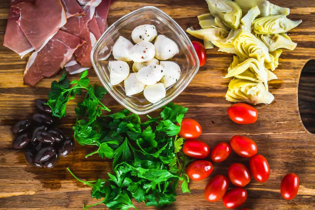 ingredients for antipasto skewers including cured meat, baby mozzarella, artichokes, cherry tomatoes, olives, and fresh herbs.