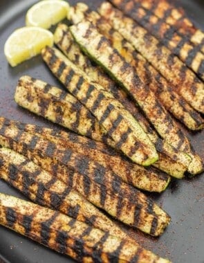 grilled zucchini on a plate, sprinkled with sumac, with lemon slices on the side.