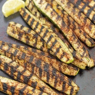 grilled zucchini on a plate, sprinkled with sumac, with lemon slices on the side.