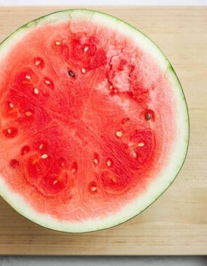 half a watermelon sitting flesh side up on a cutting board next to a large knife.