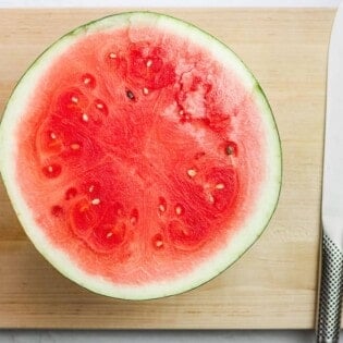 half a watermelon sitting flesh side up on a cutting board next to a large knife.