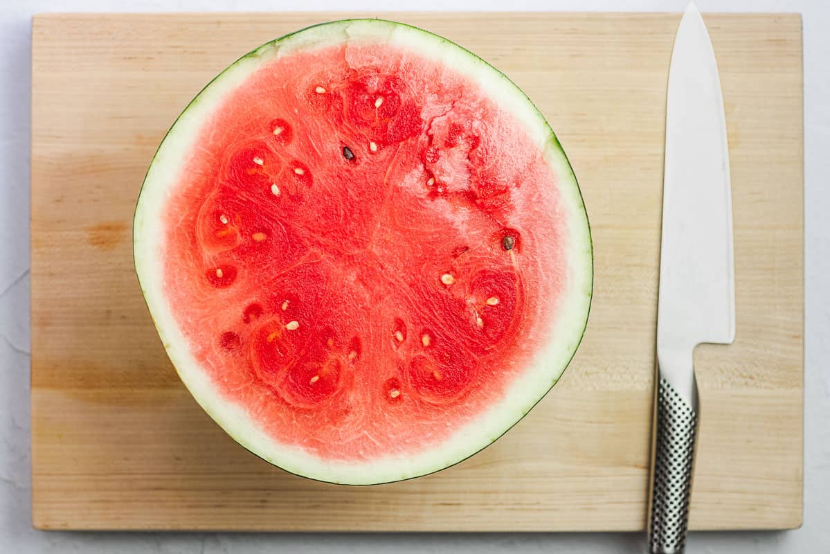 half a watermelon sitting flesh side up on a cutting board next to a large knife.