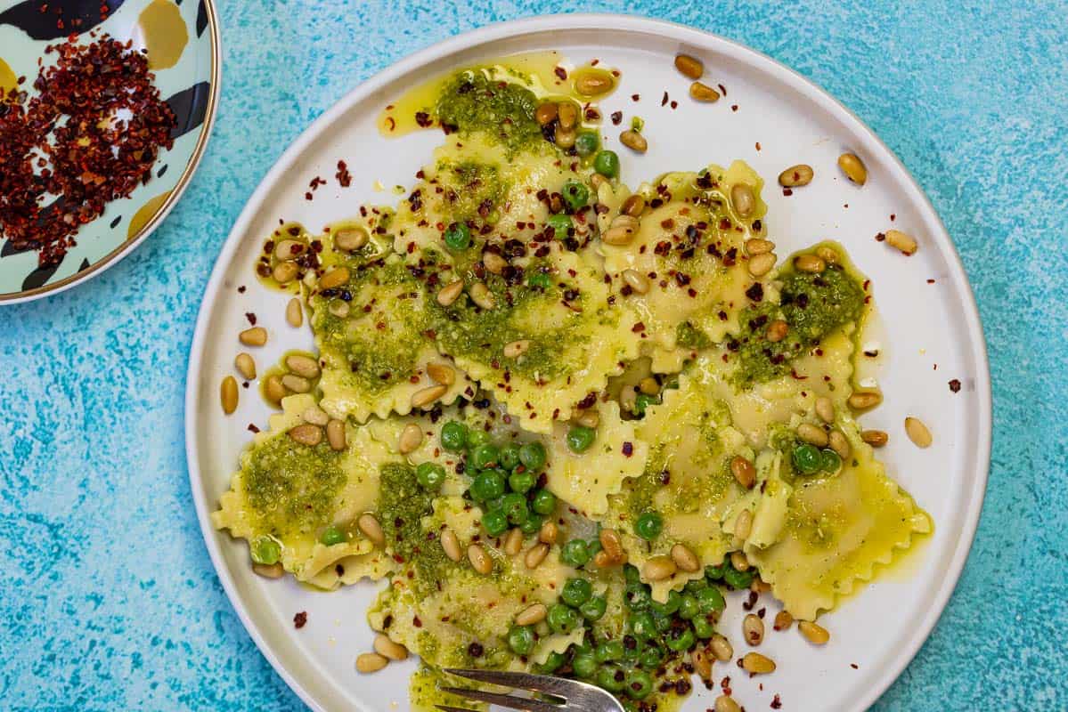 pesto ravioli and peas on a plate with a sprinkle of Aleppo pepper and toasted pine nuts on top.leppo pepper