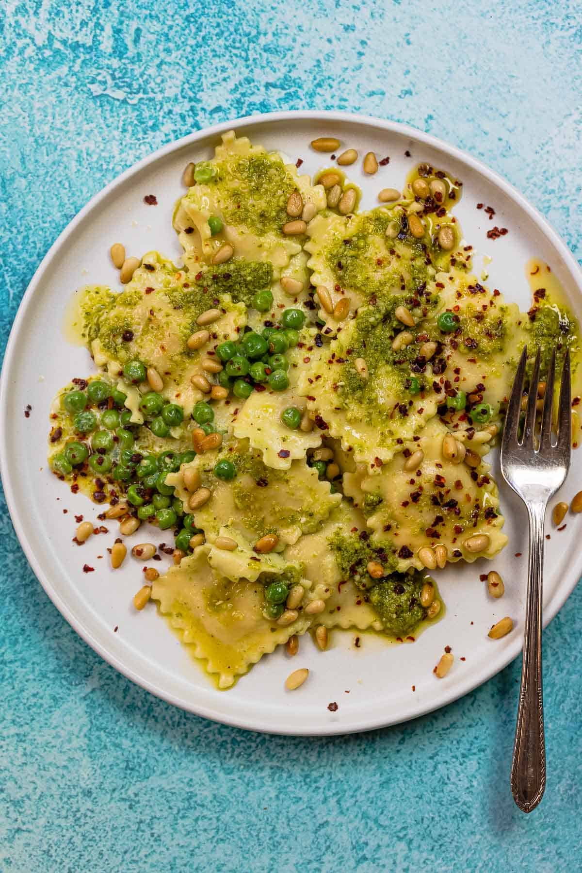 pesto ravioli recipe with peas, toasted pine nuts, and Aleppo pepper flakes on a plate with a fork.