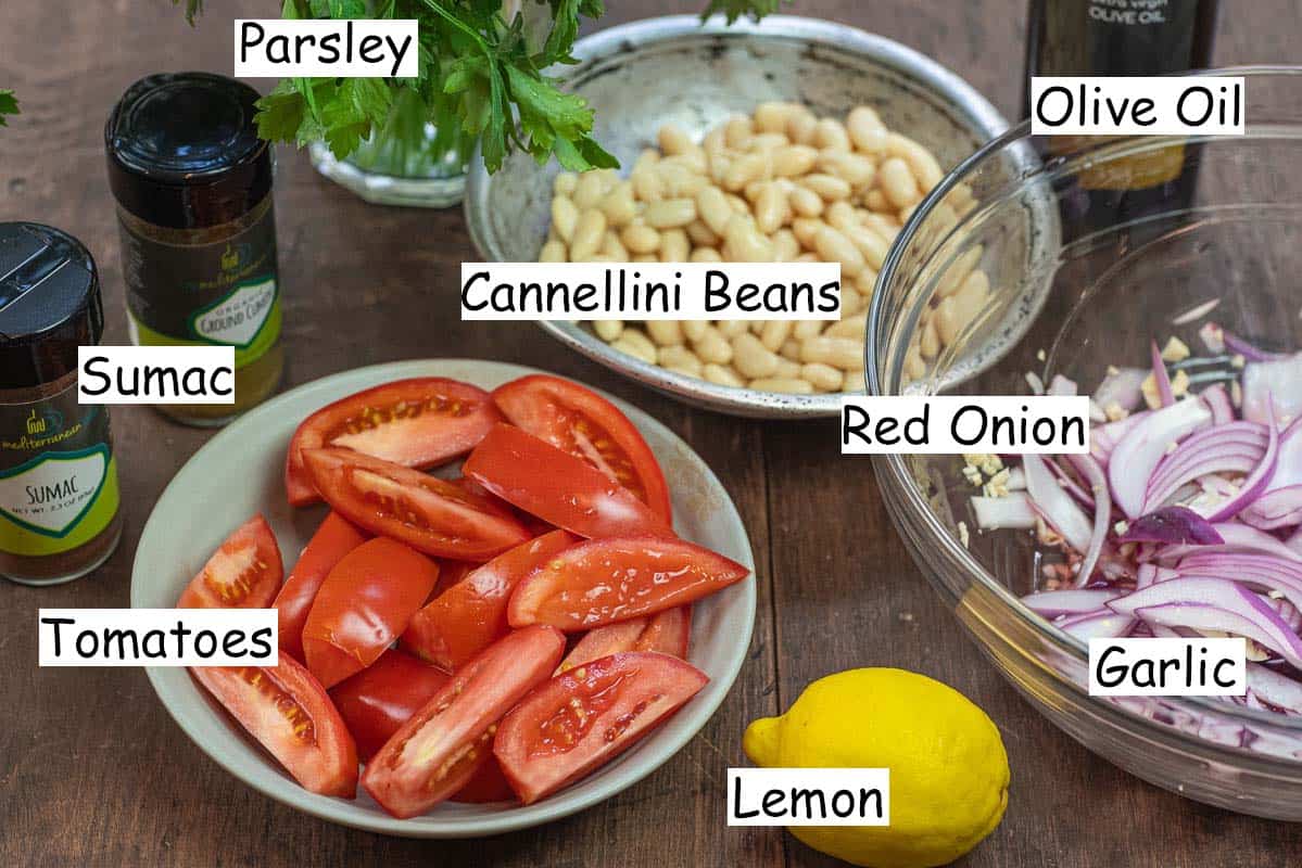 labeled ingredients for piyaz including parsley, sumac, ground cumin, tomatoes, lemon, garlic, red onion, cannellini beans, and olive oil.