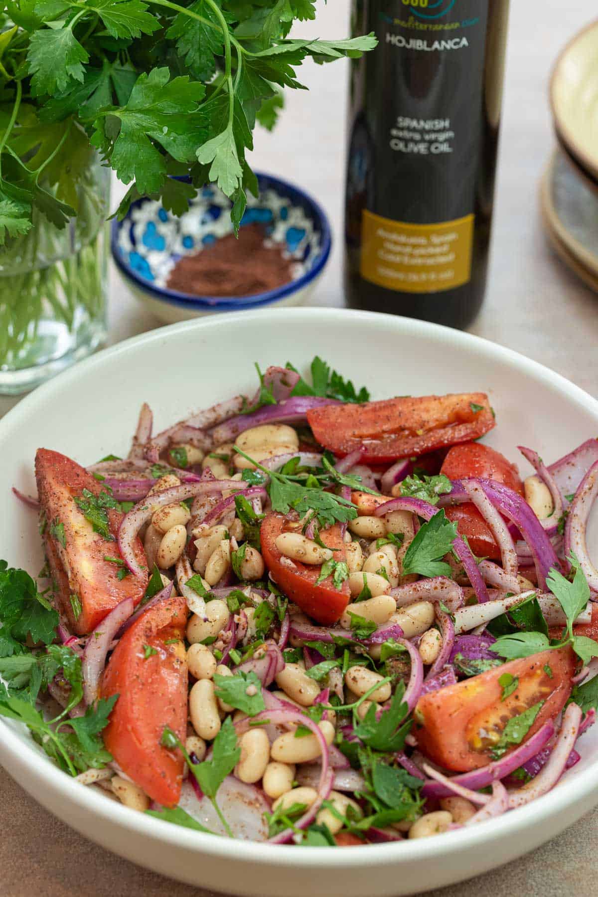 Turkish bean salad in a white bowl with fresh parsley, sumac, and olive oil in the background.