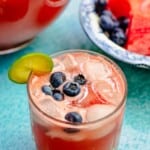 watermelon sangria in a glass with ice and blueberries, with a lime slice on the glass. Fruit and more sangria in the background.