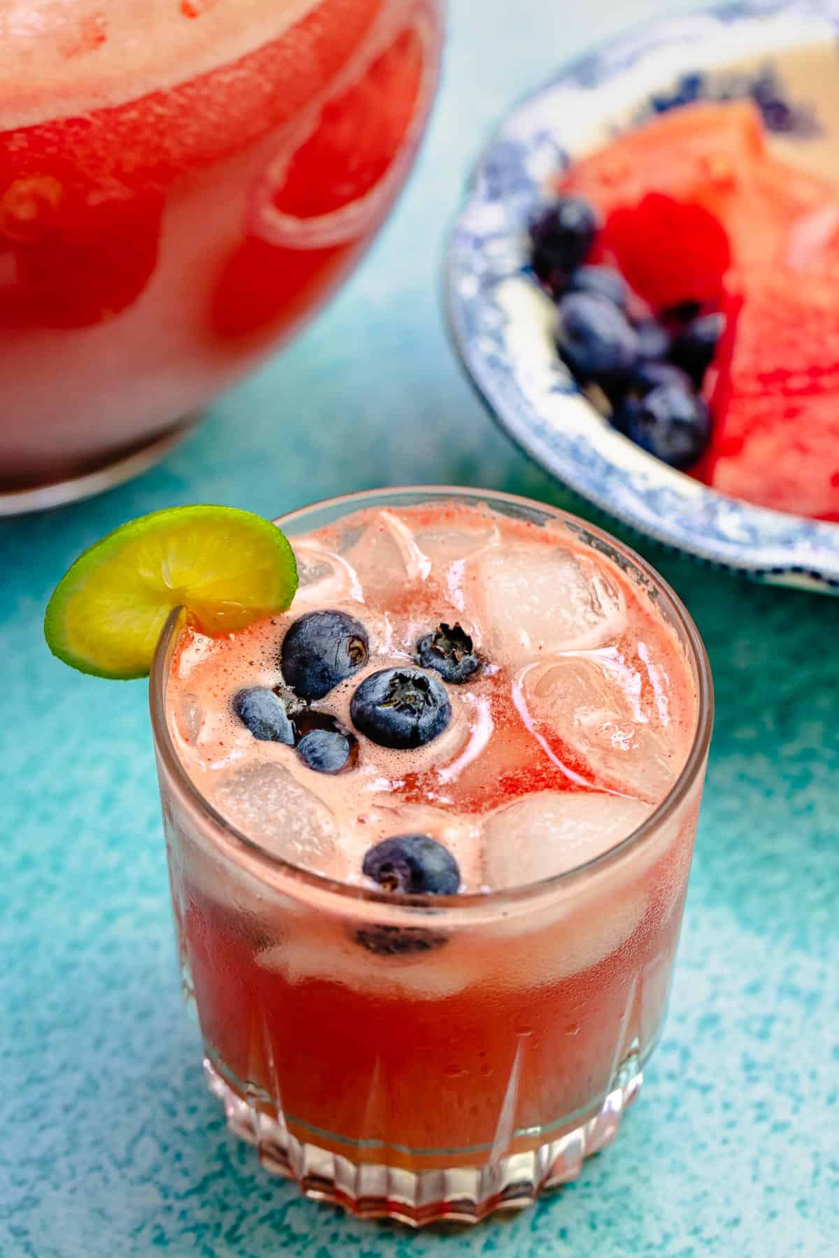 watermelon sangria in a glass with ice and blueberries, with a lime slice on the glass. Fruit and more sangria in the background.