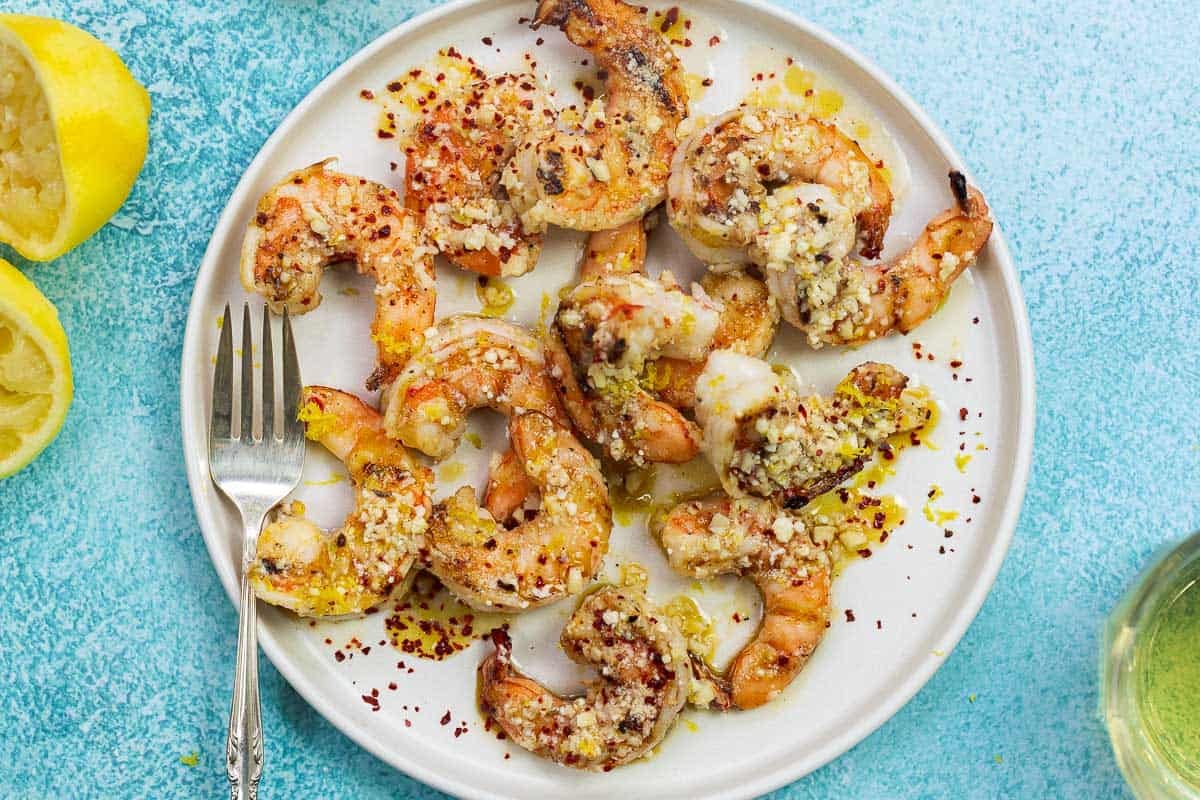 grilled jumbo shrimp with sauce and Parmesan cheese on a white plate with lemon halves in the background.