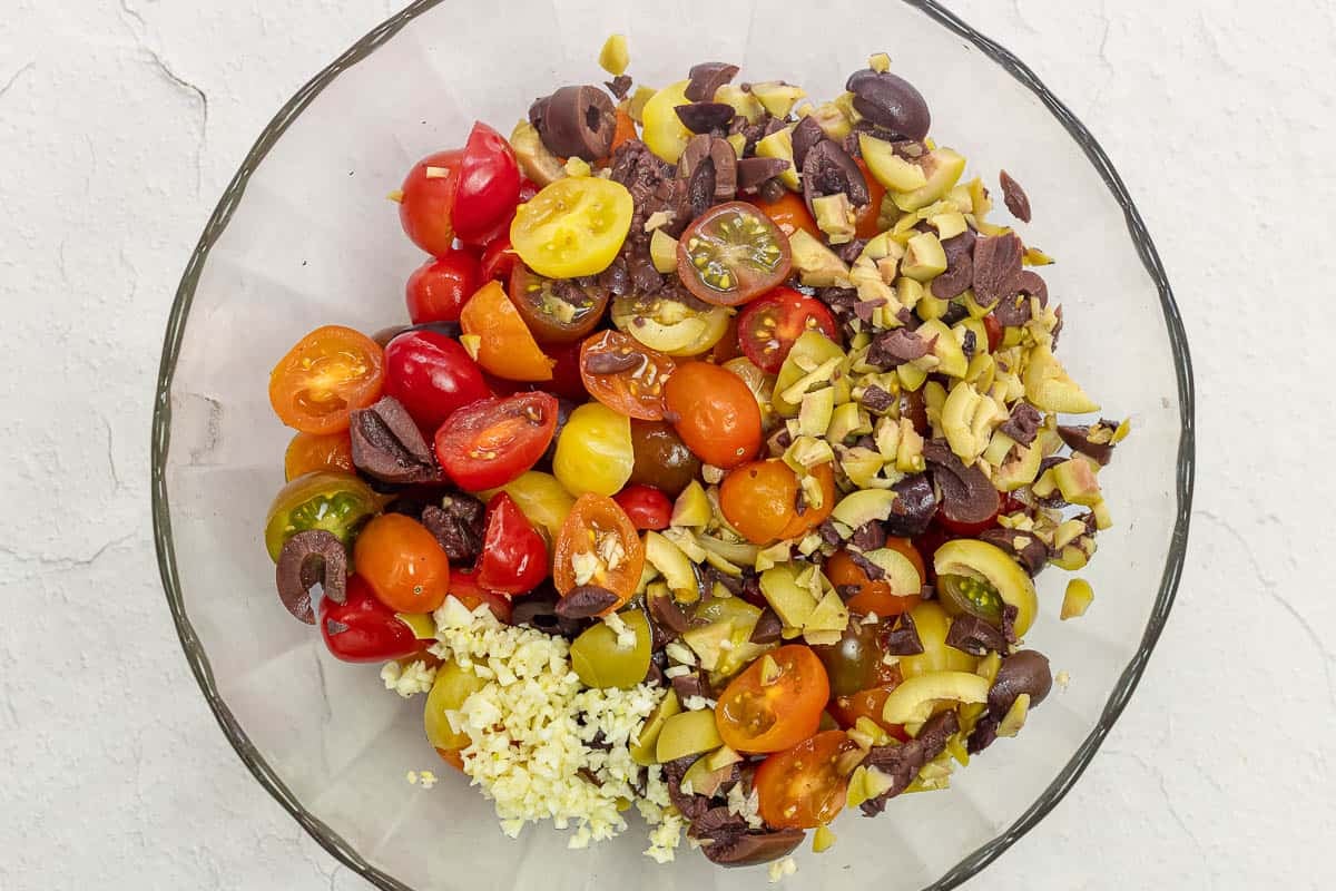 ingredients for cherry tomato salad in a bowl, including sliced tomatoes, chopped olives, and garlic.