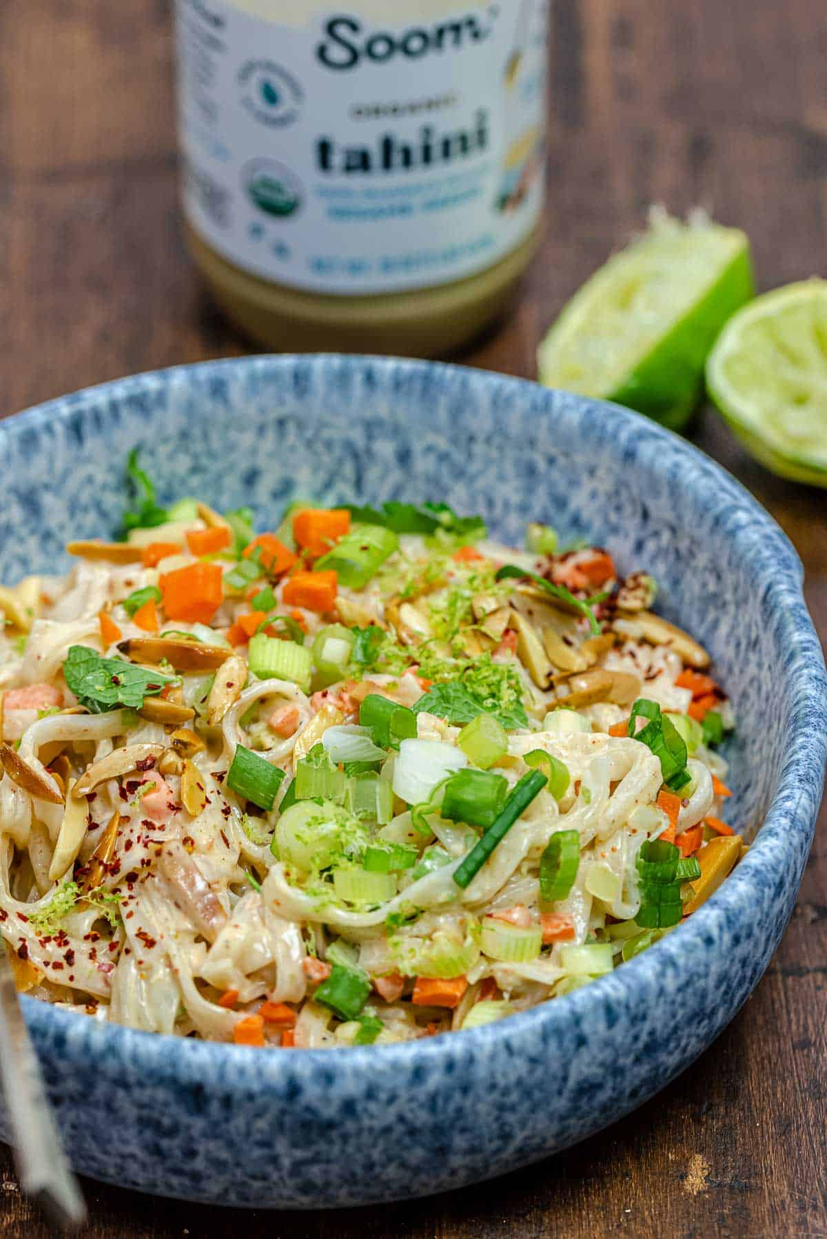 cold noodle salad with tahini dressing and vegetables in a bowl with a jar of tahini and juiced limes in the background.