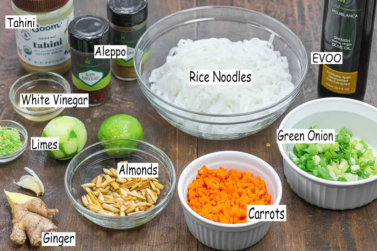labeled ingredients for noodle salad including rice noodles, extra virgin olive oil, green onions, carrots, slivered almonds, ginger, garlic, limes and lime zest, tahini, white vinegar, tahini, Aleppo pepper, and ground cumin