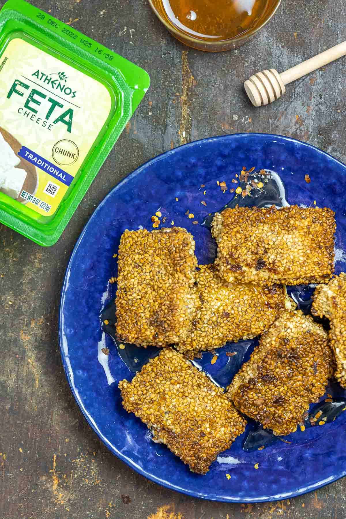 feta saganaki with sesame seeds, with a block of feta cheese and a bowl of honey to the side.