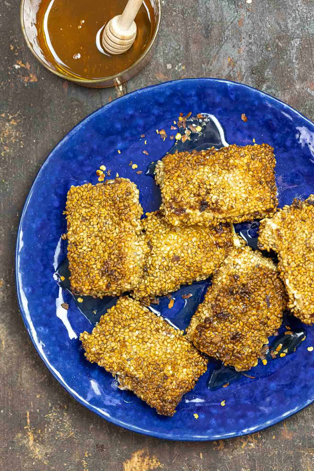 fried feta cheese on a blue plate with a bowl of honey to the side.