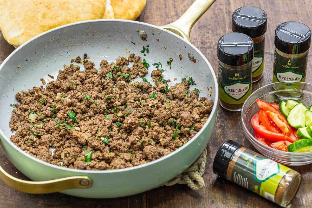 ground beef in a skillet with spices from The Mediterranean Dish next to it. There is also a small bowl with sliced cucumber and tomatoes.
