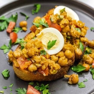 chickpeas on toast with eggs on top on a dark plate.