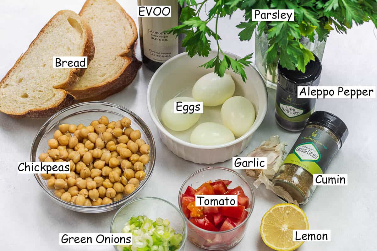 labeled ingredients for smashed chickpea toast including chickpeas, bread, olive oil, parsley, Aleppo pepper, ground cumin, lemon, garlic, tomato, green onions, and boiled eggs.
