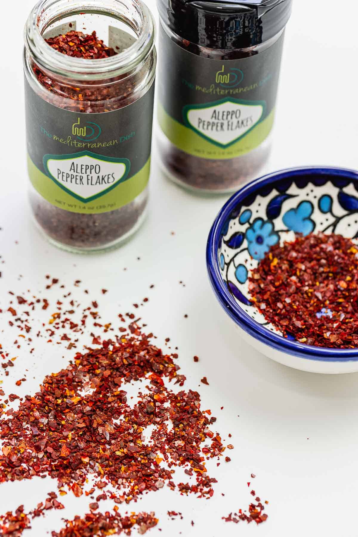 Two bottles of the Mediterranean Dish Aleppo Pepper Flakes, as well as some in a small bowl, and some sprinkled on a white surface.