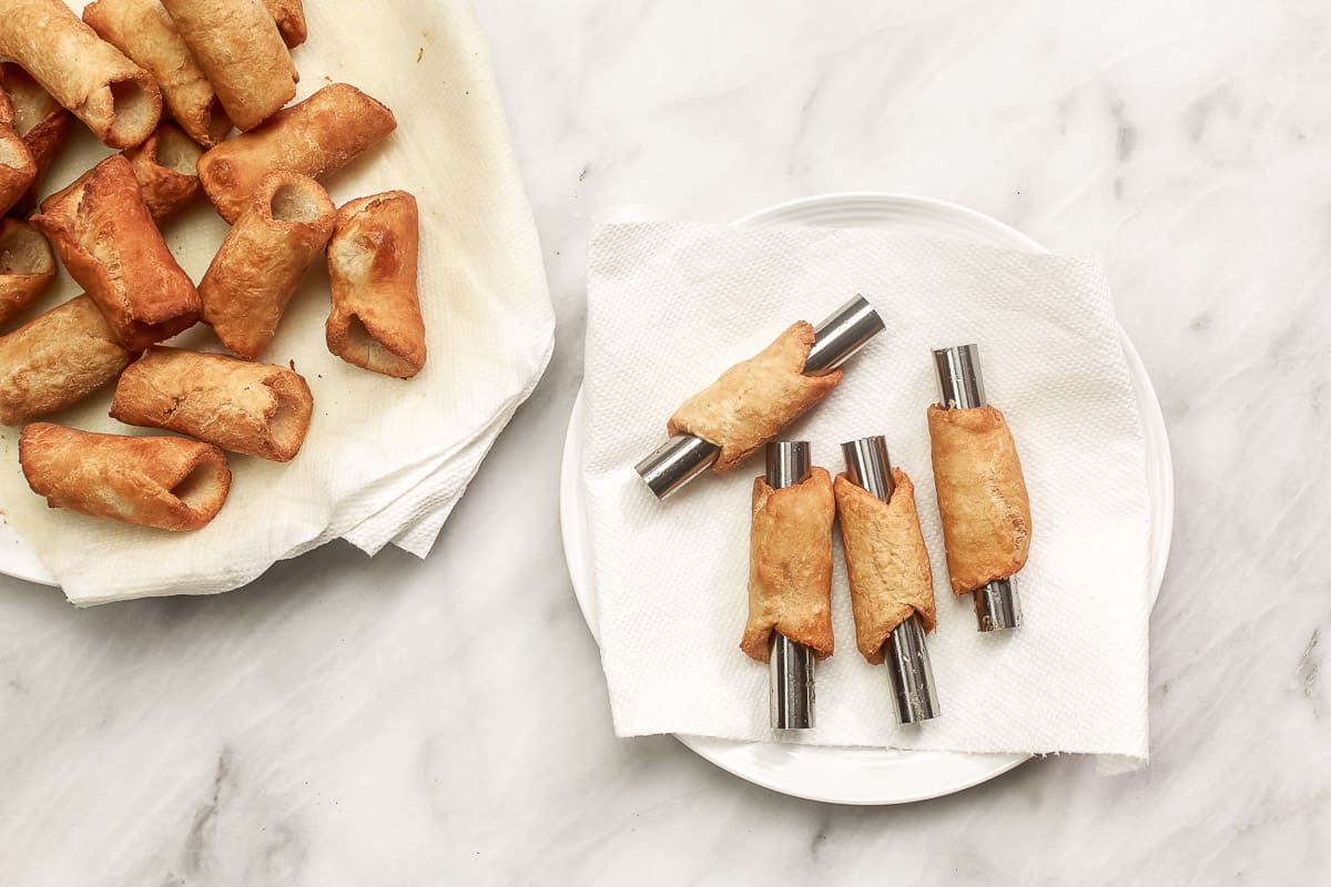 a plate with fried cannoli shells cooling paper towels, next to a plate of fried shells still on the mold.