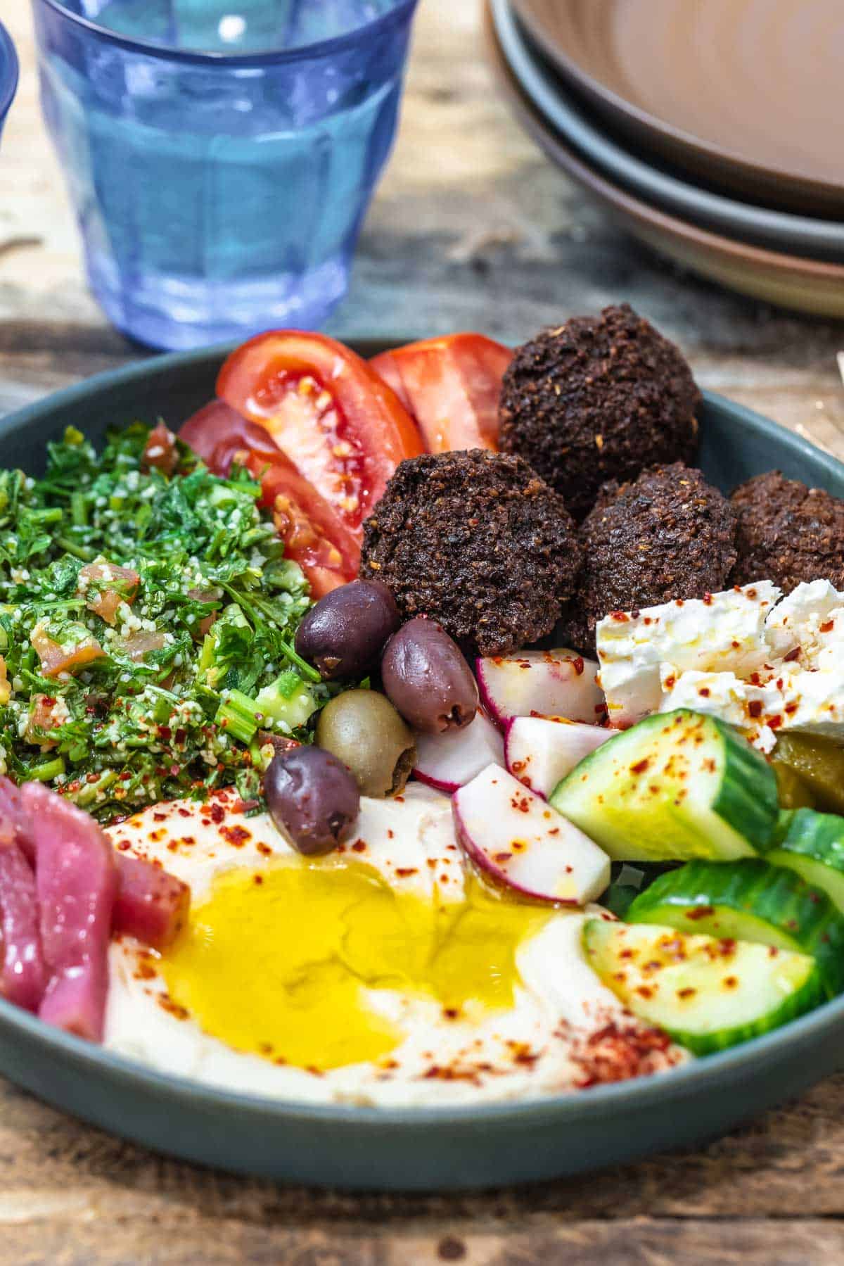 angle shot of one Mediterranean bowl of falafel with hummus, fresh vegetables, tabouli, and olive oil. A glass of water and bowls to the side