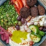 a close up of a falafel bowl assembled with hummus, pickles, olives , fresh vegetables and a tabouli salad