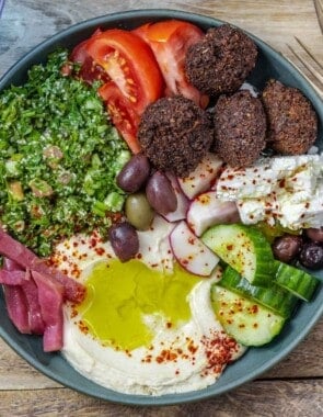 a close up of a falafel bowl assembled with hummus, pickles, olives , fresh vegetables and a tabouli salad