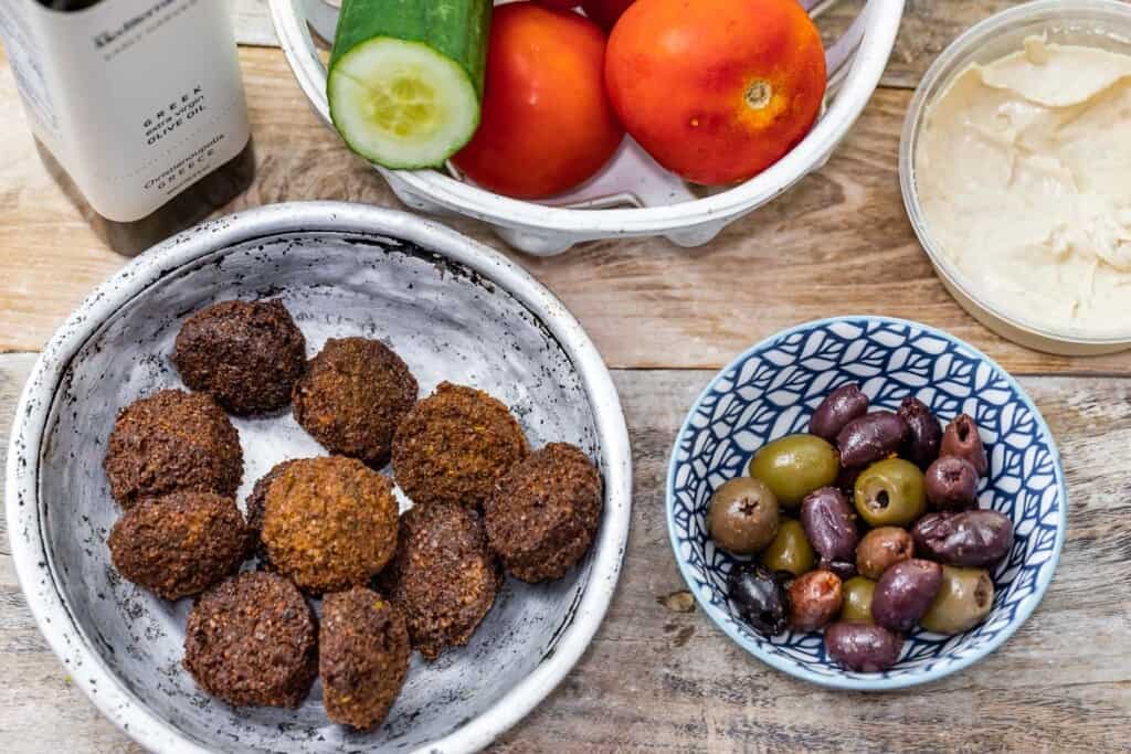 ingredients for falafel bowl including cooked falafel, olives, hummus, tomatoes, and cucumbers
