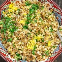 Toasted and cooked Israeli couscous in a patterned bowl with fresh herbs and lemon zest sprinkled on top.