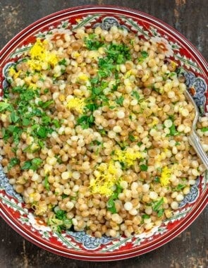 Toasted and cooked Israeli couscous in a patterned bowl with fresh herbs and lemon zest sprinkled on top.