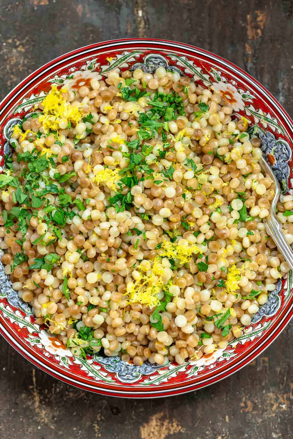 Toasted and cooked Israeli couscous in a patterned bowl with fresh herbs and lemon zest sprinkled on top.
